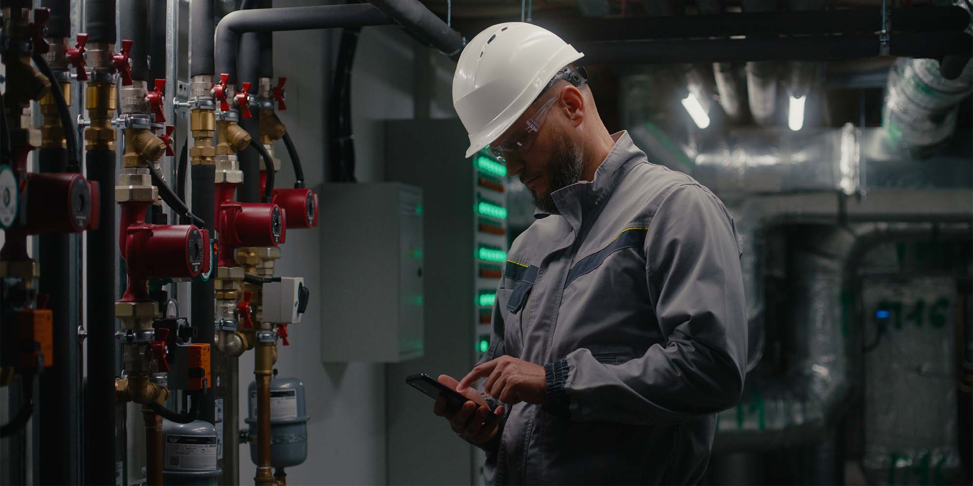 Industrial engineer in a hard hat and safety glasses using a smartphone to monitor system status in a facility with complex piping and valve systems, highlighting modern industrial workflow and remote management capabilities.